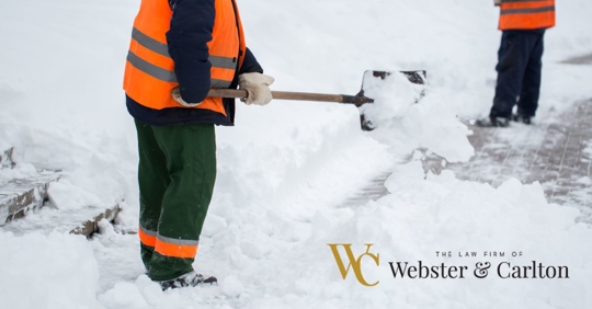 Two people in bright orange vests shoveling a snow-filled street.